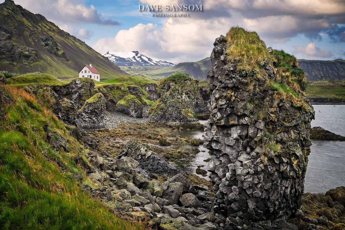 Coastal basalt cliffs and a lone white house beneath snow-capped Icelandic mountain