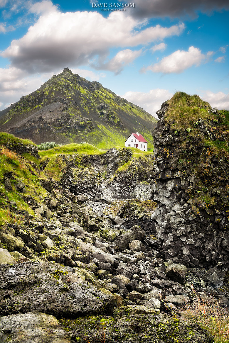 Steep green Icelandic mountain rising above basalt cliffs with a white house in the valley
