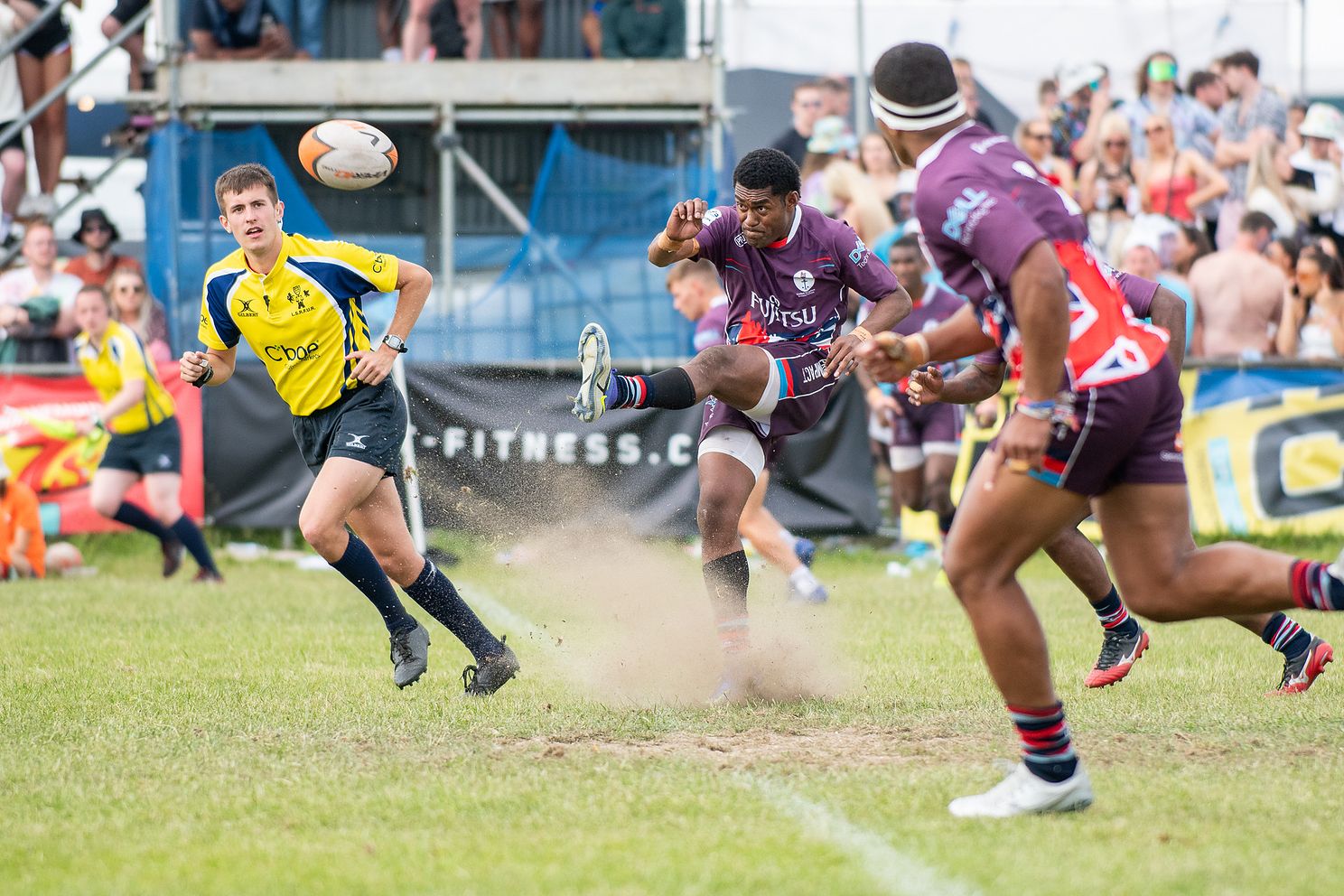 Rugby sevens action as a player kicks the ball, with a referee and a lively crowd watching