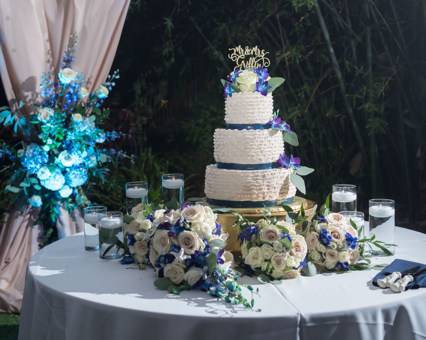 four-tiere white wedding cake with purple details on table surrounded by flower bouquets.