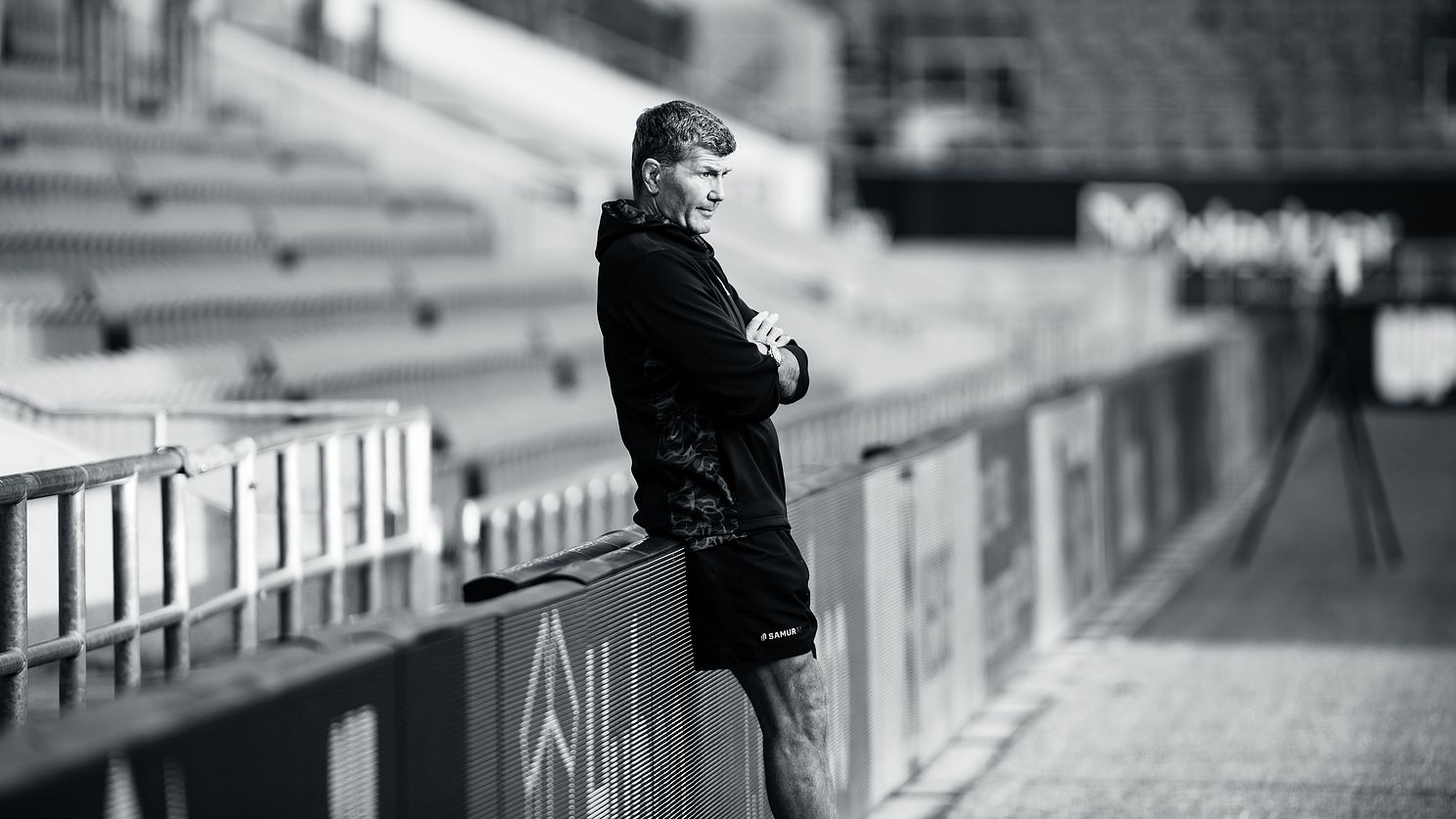 Rob Baxter of Exeter Chiefs in a black and white stadium shot, focused and leaning on a railing.
