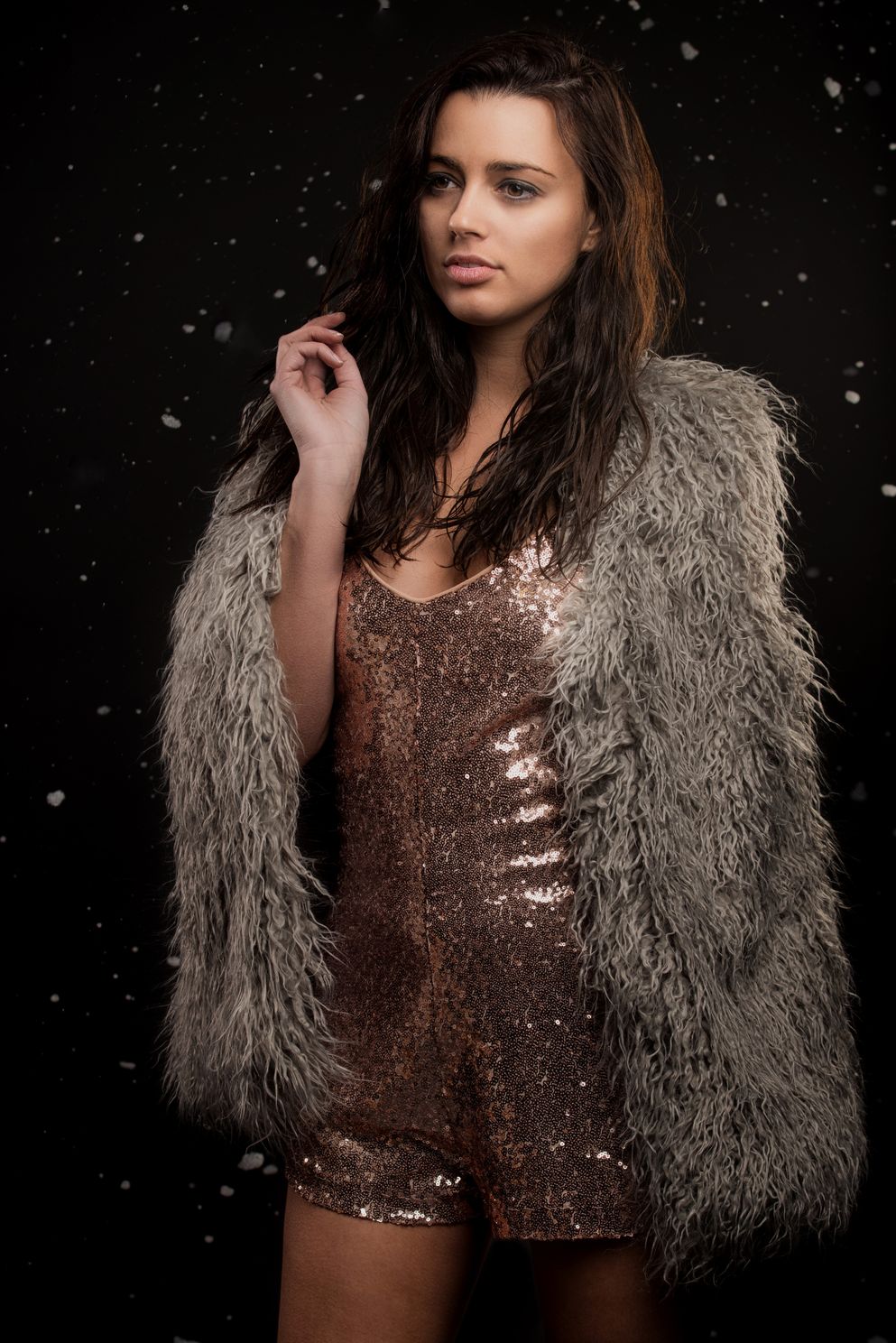 Model in studio lighting wearing fancy dress and fur coat, snow falling on a black background.