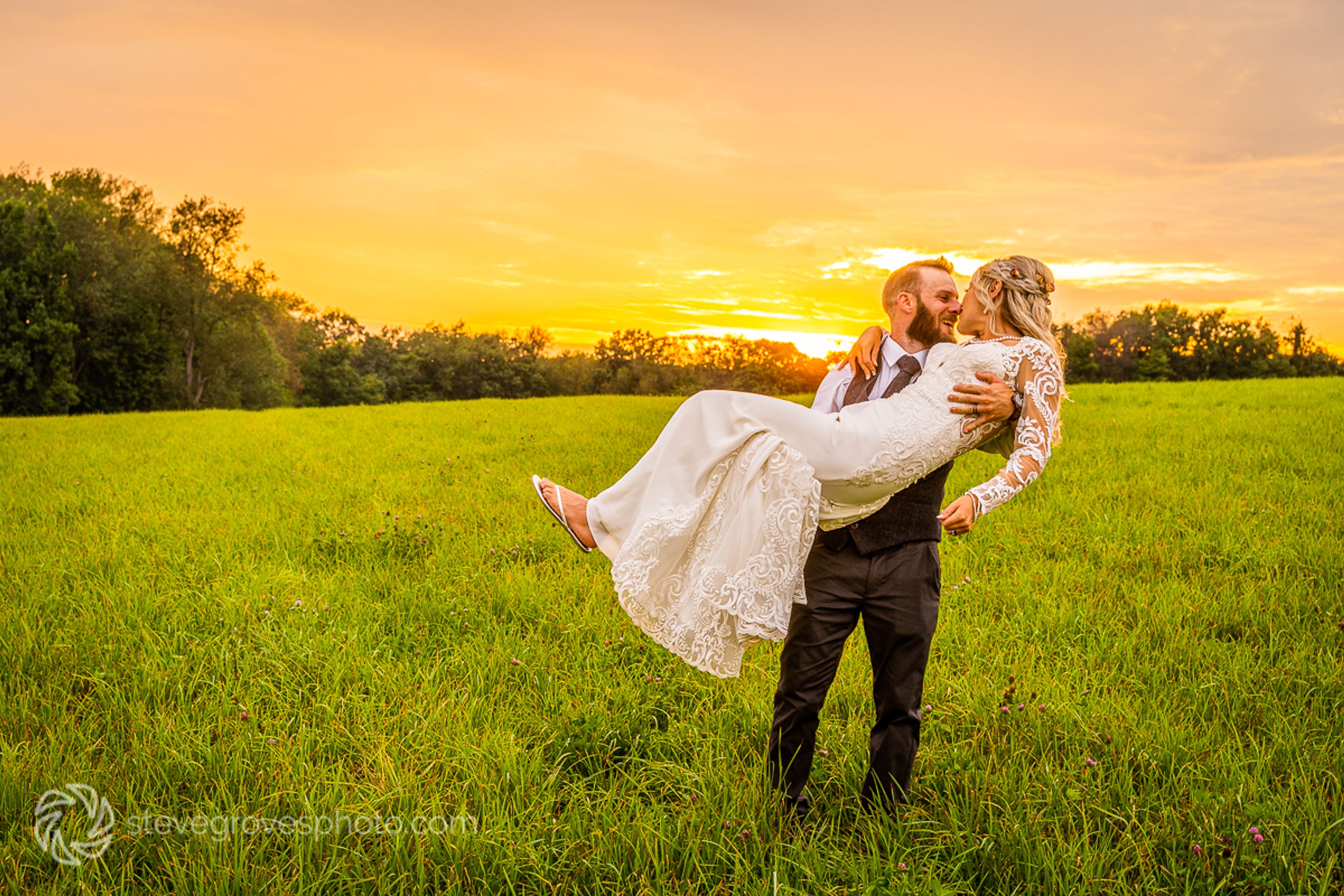 A groom holds a bride in front of a sunset.