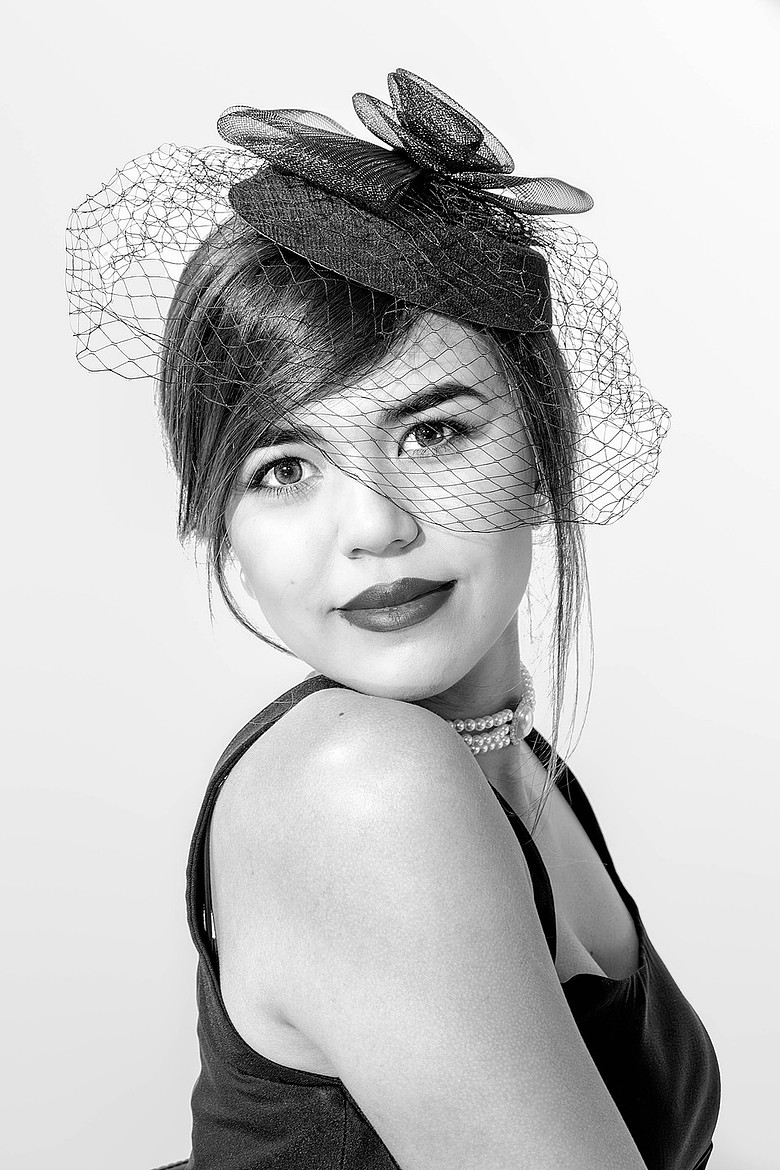 Black and white portrait of a woman in a fascinator with veil, pearl necklace, and soft expression.