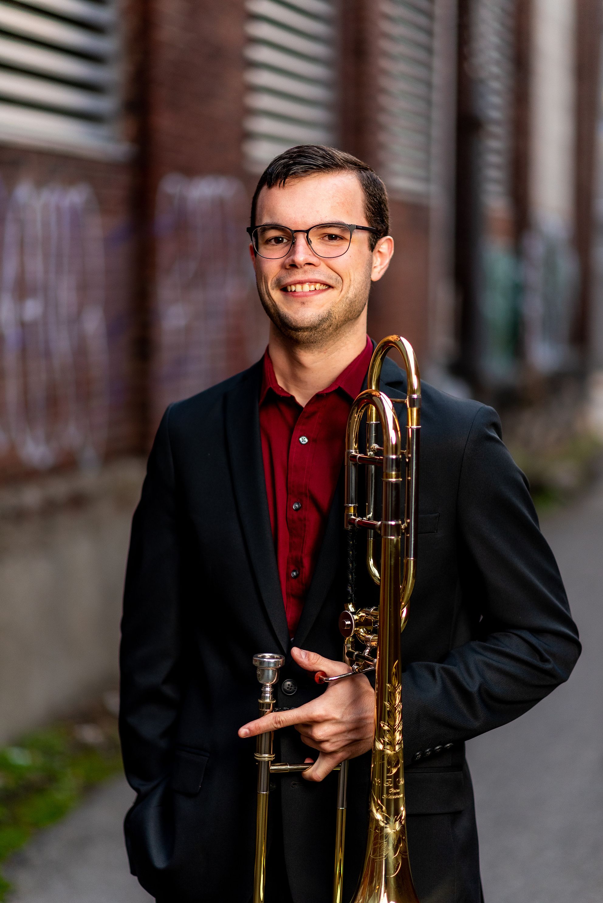 A person with a trombone poses for a photo in an alley.