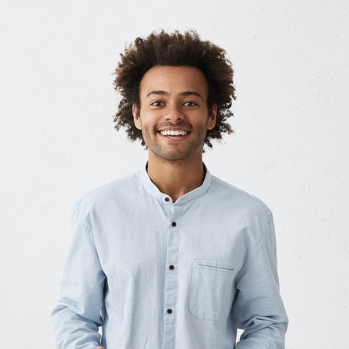 Handsome young African American male wearing light long shirt smiling and gesturing with both hands, having clueless expression on his face, standing at white wall with copy space for your text