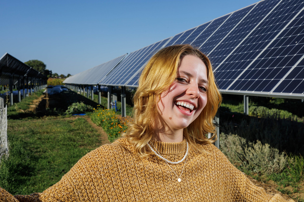 Smiling woman in a yellow sweater standing in front of solar panels on a sunny day.