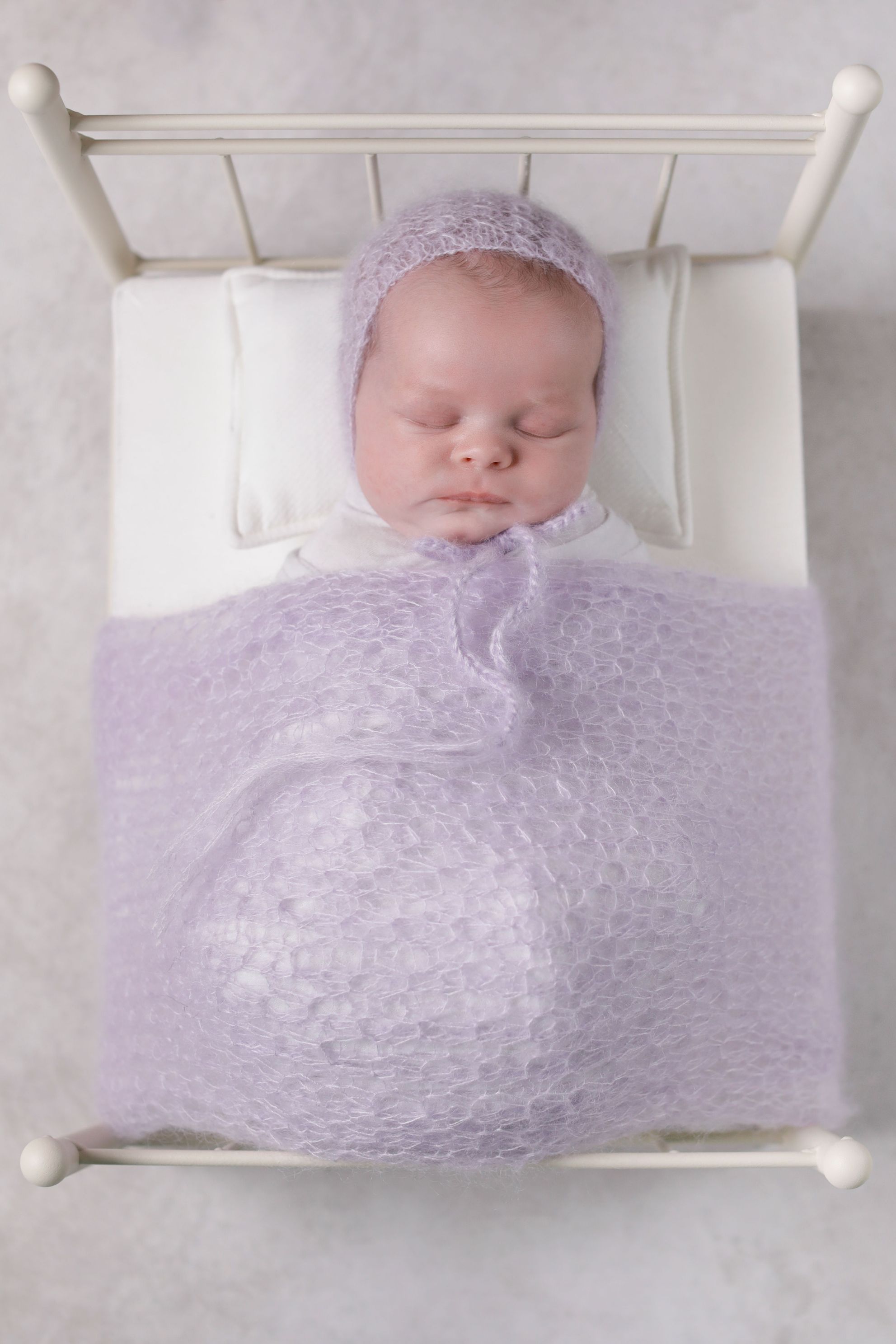White newborn bed with purple wrap with matching purple bonnet and White pillow, tucked in sleeping