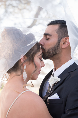 groom kissing bride's forehead with her wedding veil covering them after their ceremony.