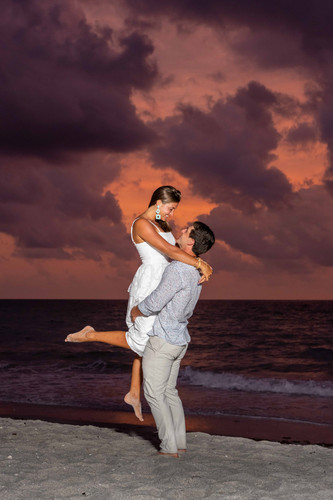 man lifting his new fiance into the air on the beach with a purple and pink sky behind them