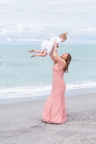 a young mother wearing a pink dress lifting her daughter in the air on the beach