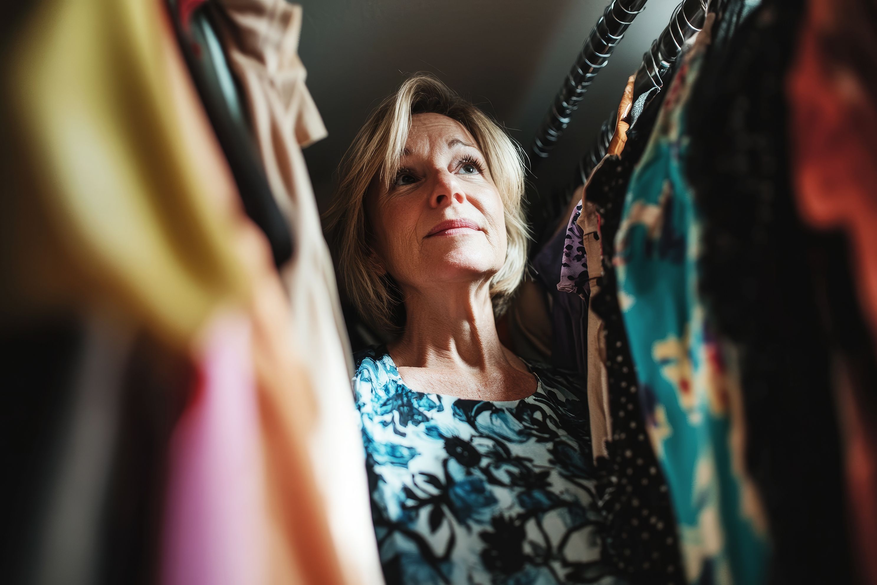 A woman stands thoughtfully in her closet surrounded by various colorful clothes.